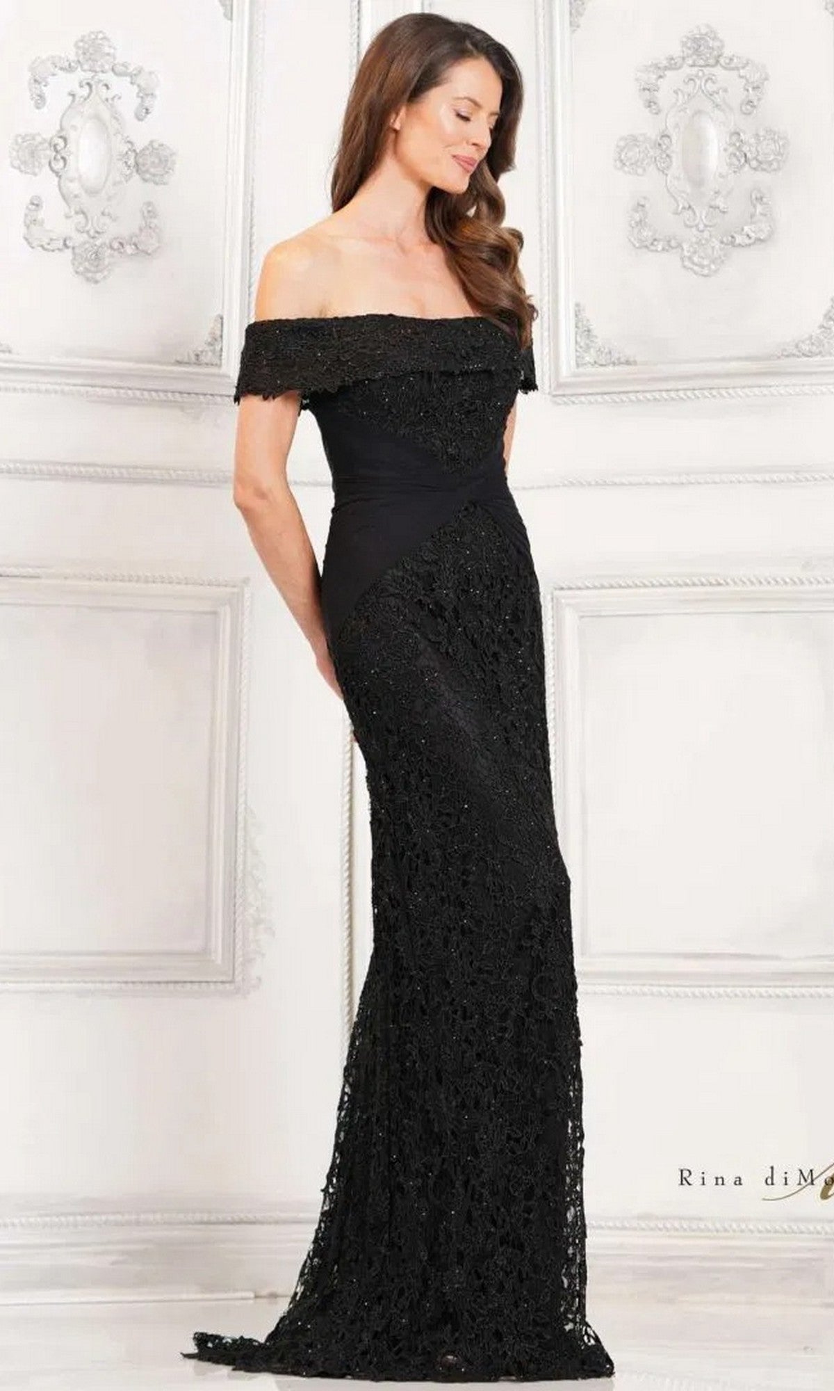 A woman in profile poses against an ornate white wall, wearing the black Long Formal Dress: Rina Di Montella RD3129, a floor-length, off-the-shoulder lace gown.